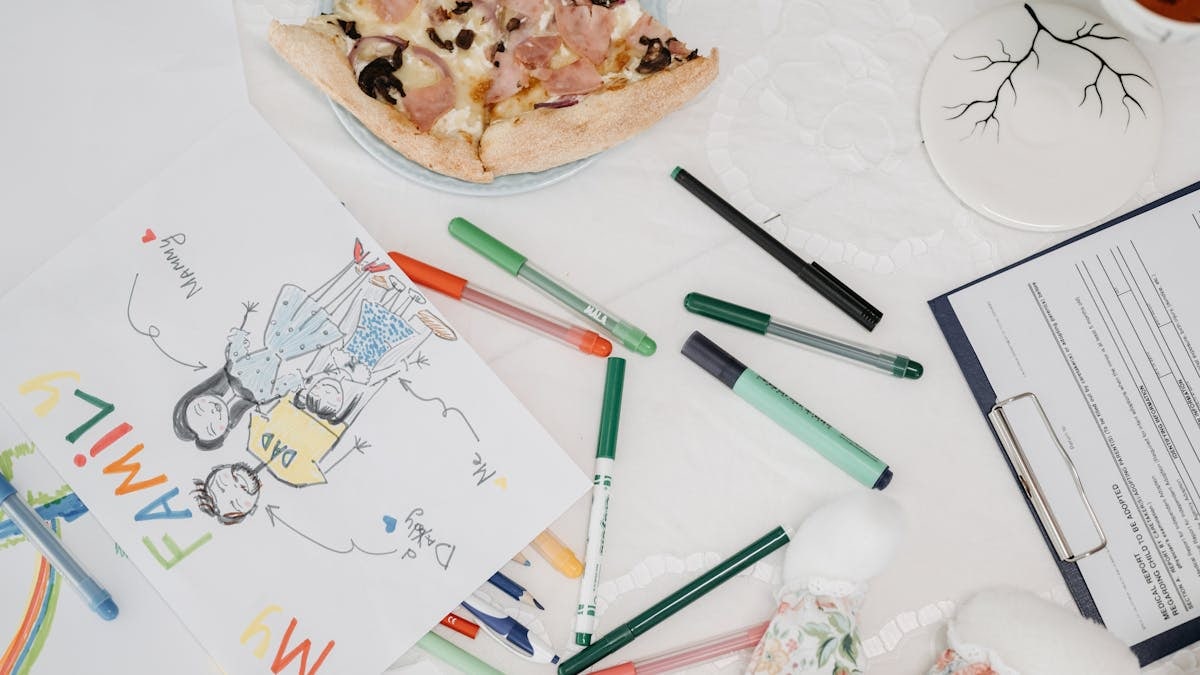 A colorful child's drawing of a family alongside pizza and pens on a table.